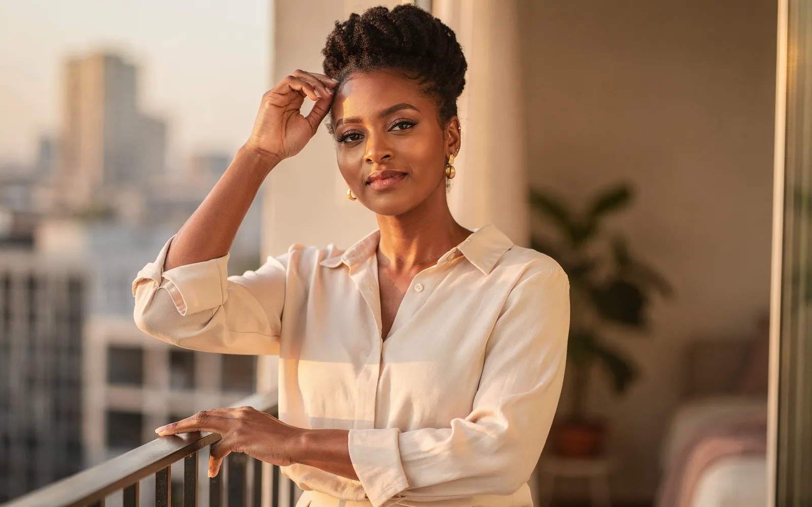 Woman with an elegant coily updo standing on a balcony at golden hour, hand resting near her hairline.