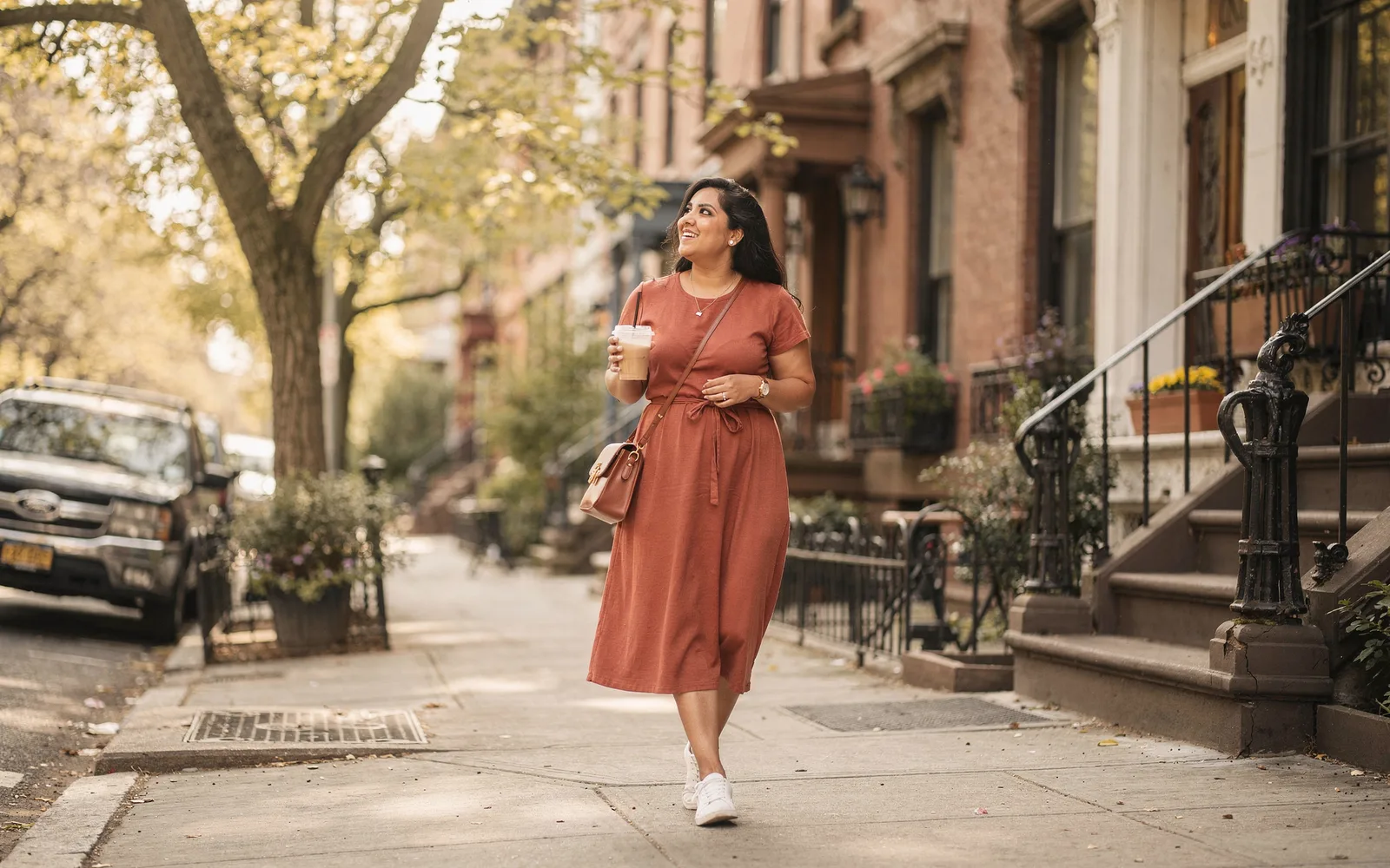A woman in a terracotta dress walks a tree-lined city street with an iced coffee, smiling slightly in golden light.