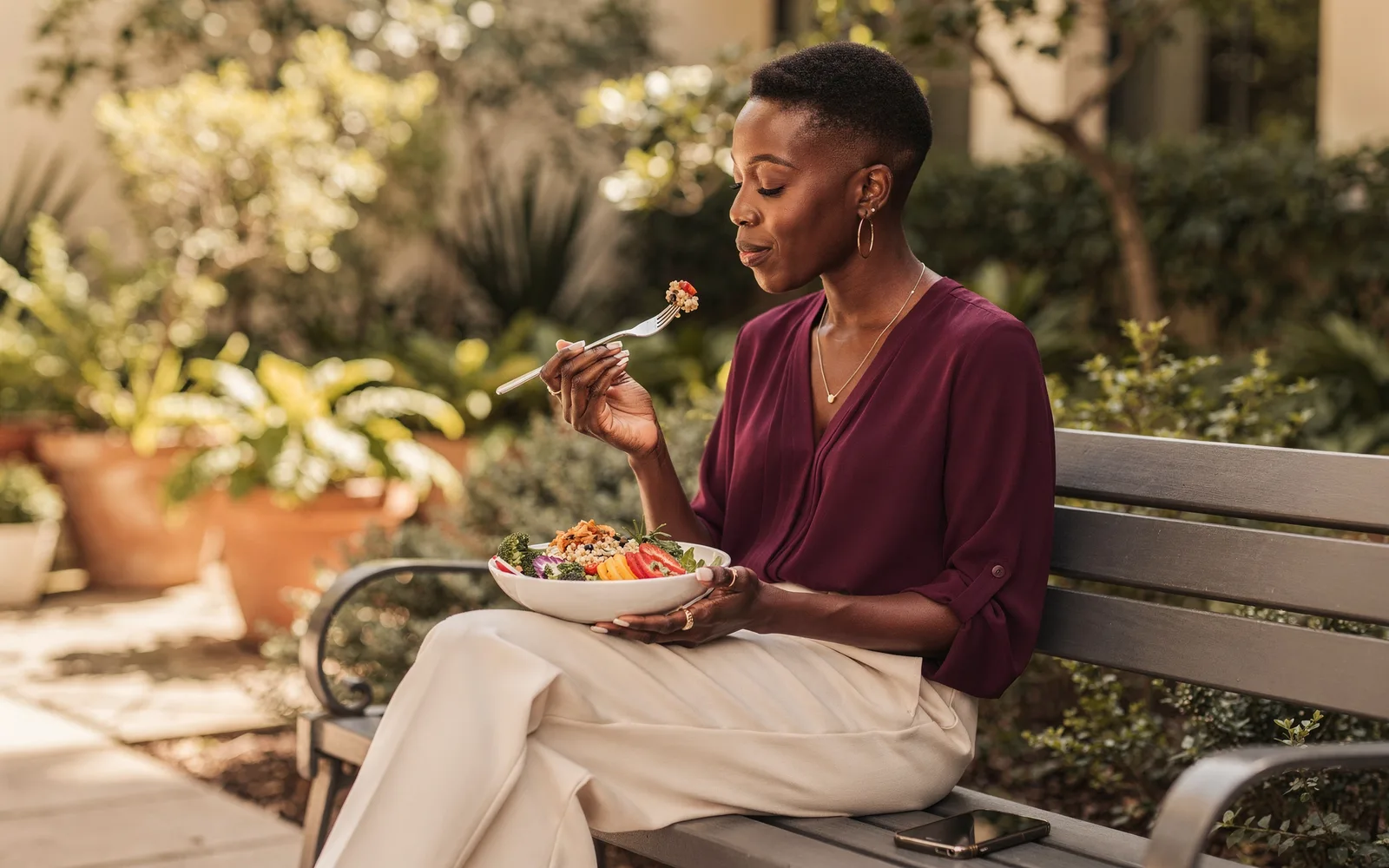 Woman enjoying a colorful grain bowl on a leafy courtyard bench at midday.