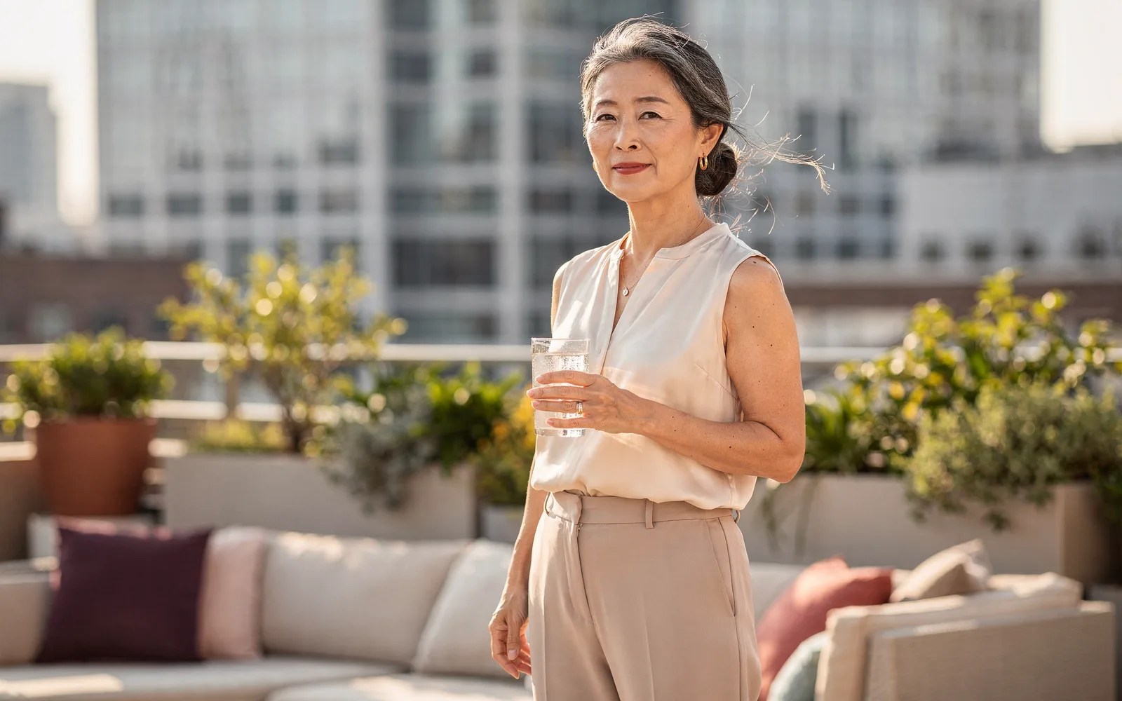 Woman in an ivory silk blouse holding iced water on a breezy planted office terrace.